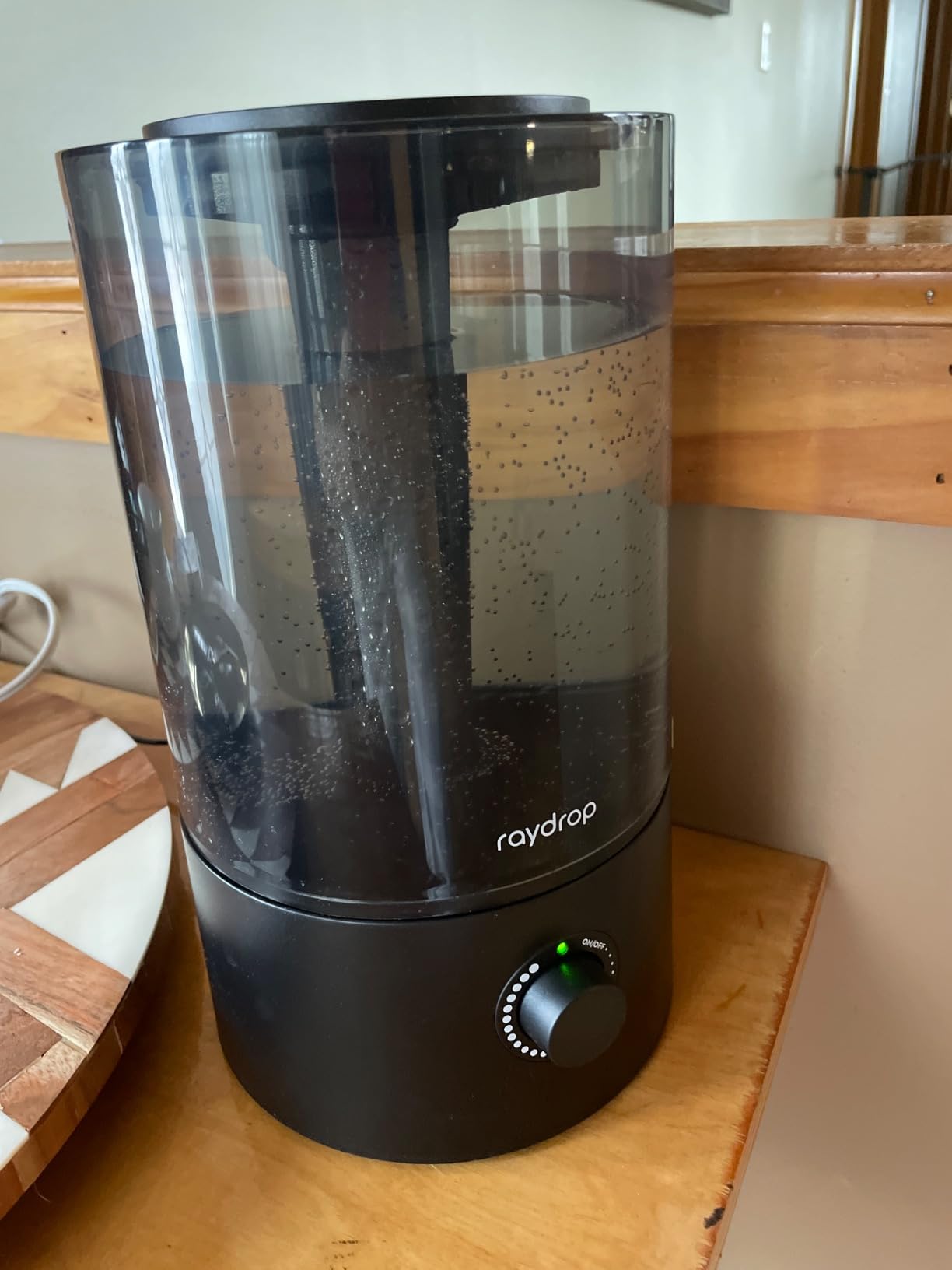 Black Raydrop humidifier with a smoked tank full of water on a wooden shelf, power light on